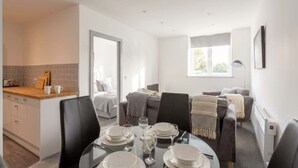 Ein moderner Essbereich mit einem Glastisch, gedeckt für vier Personen, eine Küche mit Holzarbeitsplatte und ein Wohnbereich mit einer Couch und einem Fenster.