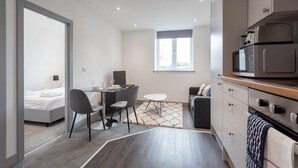 A modern hotel room with a bed, a desk with a chair, a dining table with chairs, a sofa, a kitchen area with a microwave, and a large window providing natural light. - Bright 1BR Apt in Hunslet, Near Leeds Centre (Leeds)