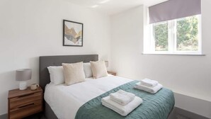 A hotel room with a bed, a nightstand, a lamp, a window with a view, and a framed picture on the wall. - Stylish 1BR Apt, Hunslet Leeds City Centre (Leeds)