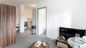 A modern living room with a sofa, coffee table, and a TV. There is a dining area with a table set for two. A trash can is visible in the corner. - Stylish 1BR Apt, Hunslet Leeds City Centre (Leeds)