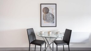 Ett modernt matområde med ett glasbord och två svarta stolar, ett inramat abstrakt konstverk på väggen och ett vitt element under.