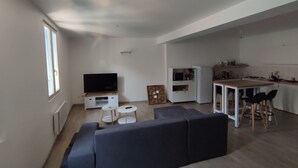 A modern living room with a large flat-screen TV, a dark-colored sofa, a small coffee table, and a dining area with a white table and chairs.