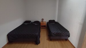 A small, narrow room with two single beds, a wooden nightstand with a lamp, and white walls.