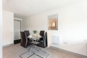 A dining area with a glass table, four black chairs, a small plant, and a mirror on the wall. - Bright & Modern 1 Bedroom Apartment in Leeds (Leeds)