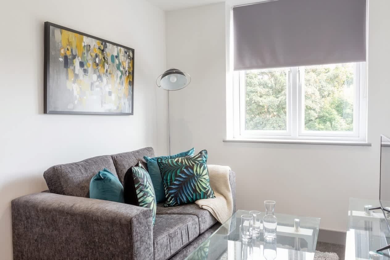 Un salon moderne avec un canapé gris, une table basse en verre et une fenêtre donnant sur un paysage verdoyant.