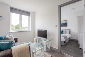 A modern hotel room with a glass coffee table, a flat-screen TV, a sofa, and a bed with a headboard.