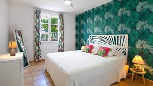 A bedroom with a bed, a ceiling fan, a window with curtains, and a wall with a leaf pattern. - Blue Bay Apartment, Orient Bay, near the beach (Collectivité de Saint-Martin)