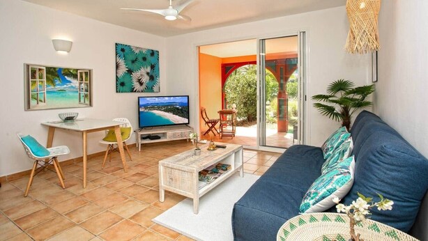 A living room with a TV, a ceiling fan, a sofa, a dining table with chairs, and a view through a glass door to an outdoor area. - Blue Bay Apartment, Orient Bay, near the beach (Collectivité de Saint-Martin)