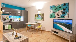 A compact living space with a kitchenette, dining area, and a TV displaying a beach scene. - Blue Bay Apartment, Orient Bay, near the beach (Collectivité de Saint-Martin)