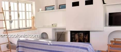 Un dormitorio con techo inclinado, una cama con colcha a rayas, una silla de fibras naturales y una chimenea.