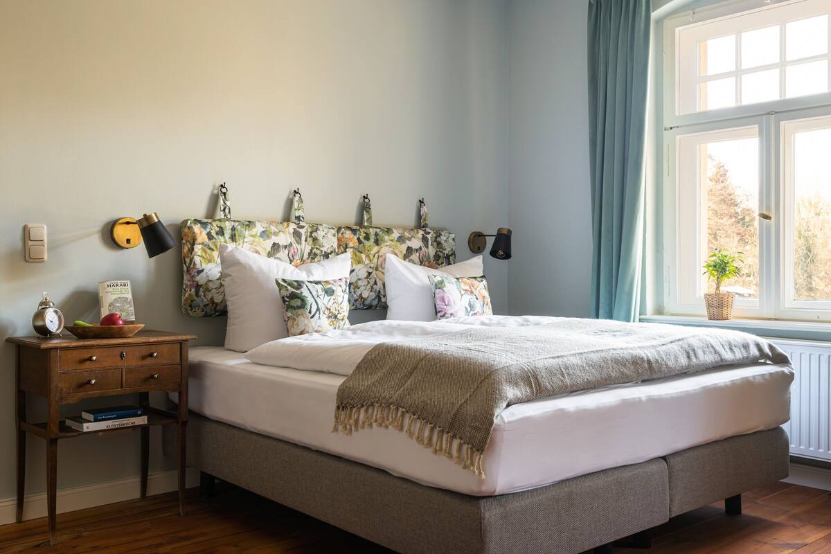 A bedroom with a bed, a bedside table with a lamp, a wall clock, and a window with curtains.
