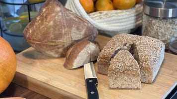 Una hogaza de pan sobre una tabla de madera con un cuchillo, un cuenco de naranjas y frascos al fondo.