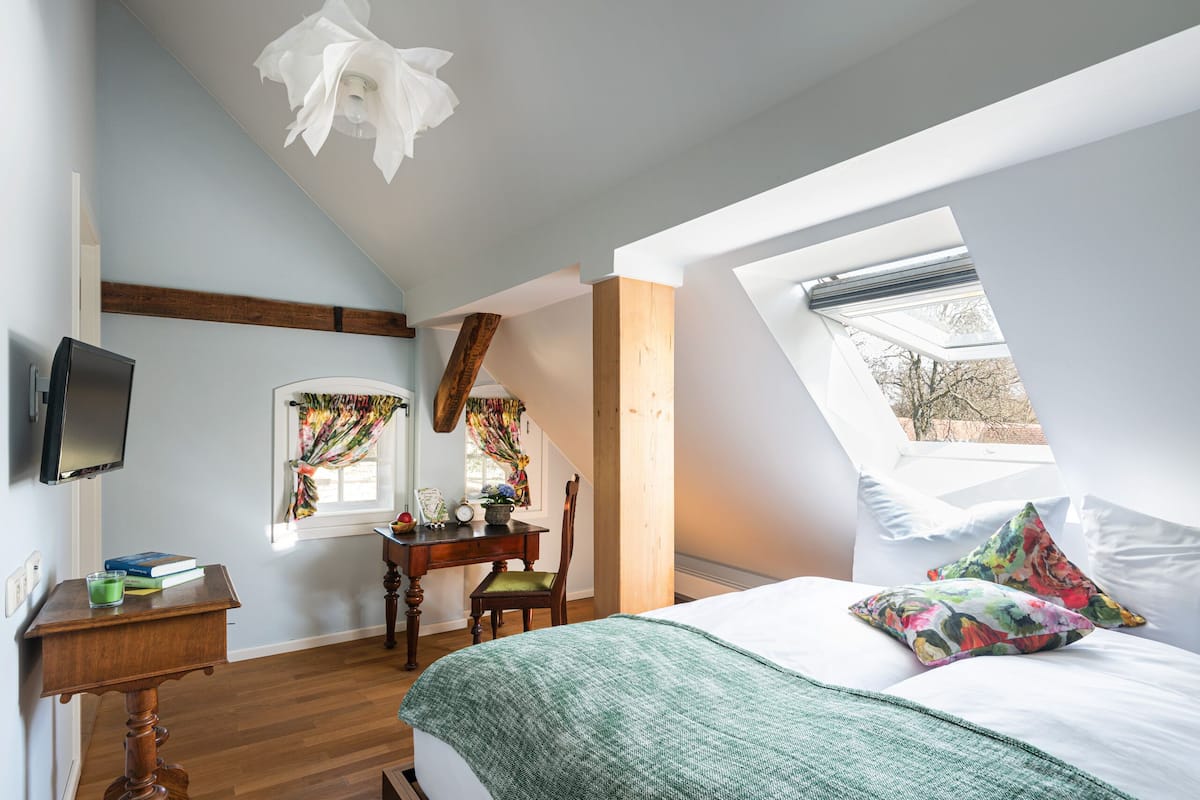 A bedroom with a sloped ceiling, a bed with white linens, a green throw blanket, a wooden desk with a chair, a flat-screen TV, and a window with curtains.
