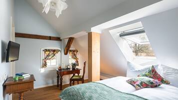A bedroom with a sloped ceiling, a bed with white linens, a green throw blanket, a wooden desk with a chair, a flat-screen TV, and a window with curtains.