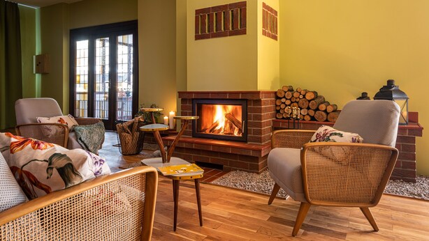 A cozy living room with a fireplace, wicker furniture, and a stack of firewood. - Klostergartenhotel Marienfliess (Marienfließ)