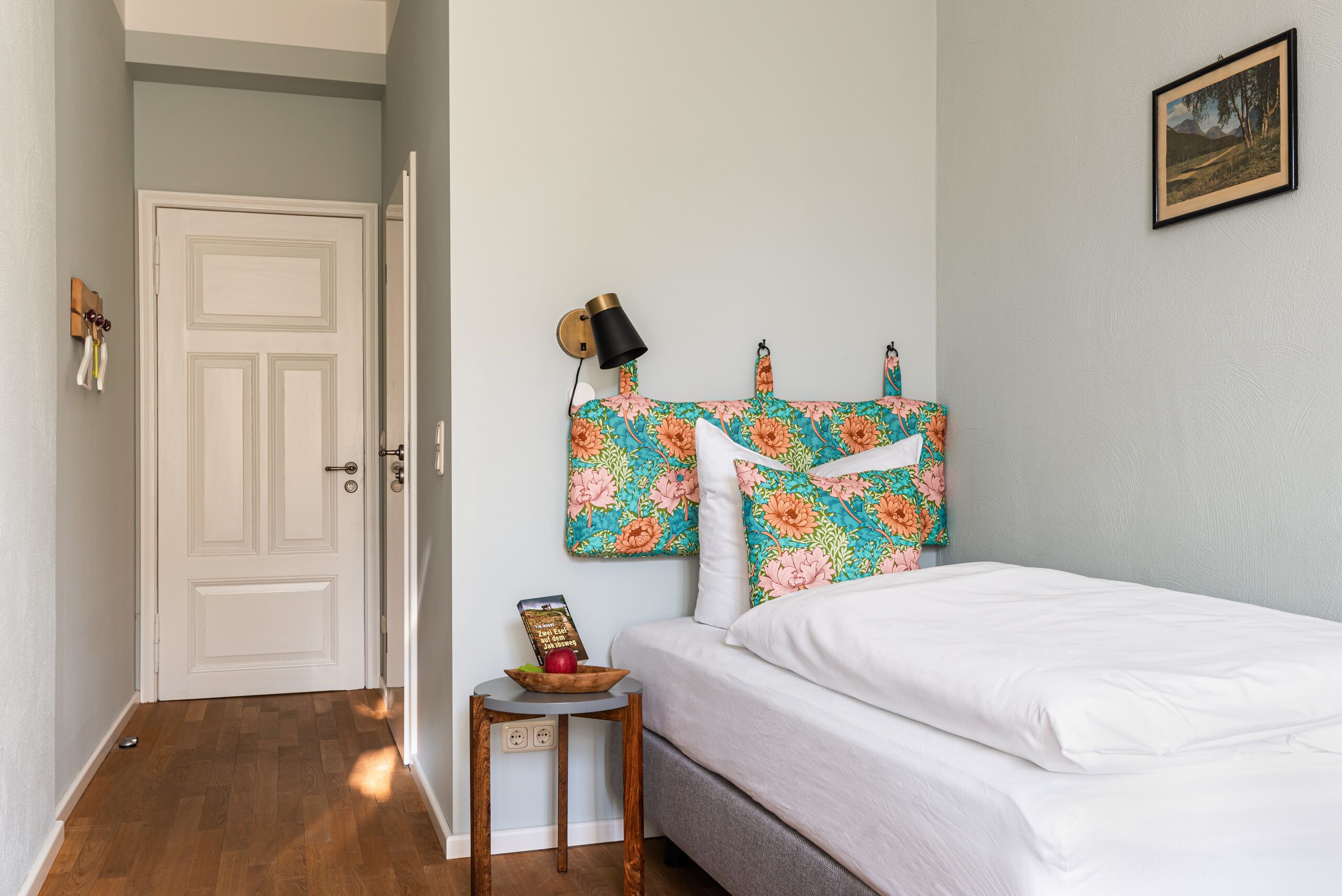 A bedroom with a bed, a nightstand with a lamp, a framed picture on the wall, and a door leading to another room.
