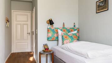 A bedroom with a bed, a nightstand with a lamp, a framed picture on the wall, and a door leading to another room.