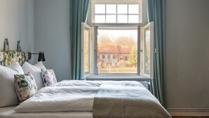 A bedroom with a large window, a bed with white bedding, and a view of a building outside. - Klostergartenhotel Marienfliess (Marienfließ)