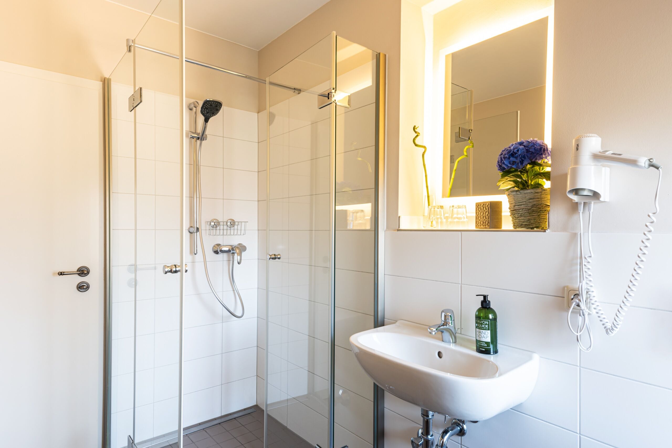 Un baño moderno con ducha de vidrio, lavamanos blanco y espejo con un florero.