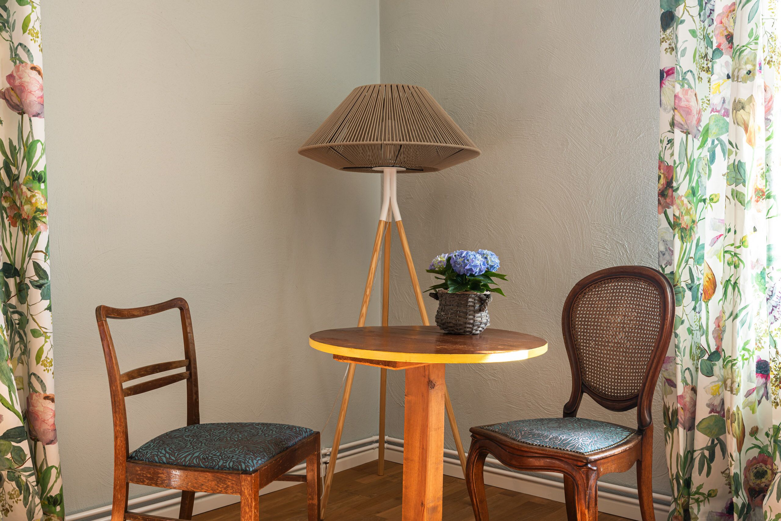 Una habitación en esquina con suelo de madera, una mesa redonda de madera con una lámpara, dos sillas y un florero con flores.