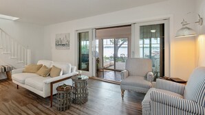Un salon spacieux avec un canapé blanc, des fauteuils à rayures et un plancher en bois. Une grande porte-fenêtre coulissante donne accès à un espace extérieur avec vue sur une étendue d’eau.