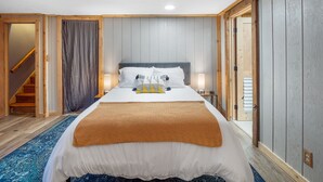 A bedroom with a bed, bedside tables, a lamp, a wooden staircase, and a view of the outside.
