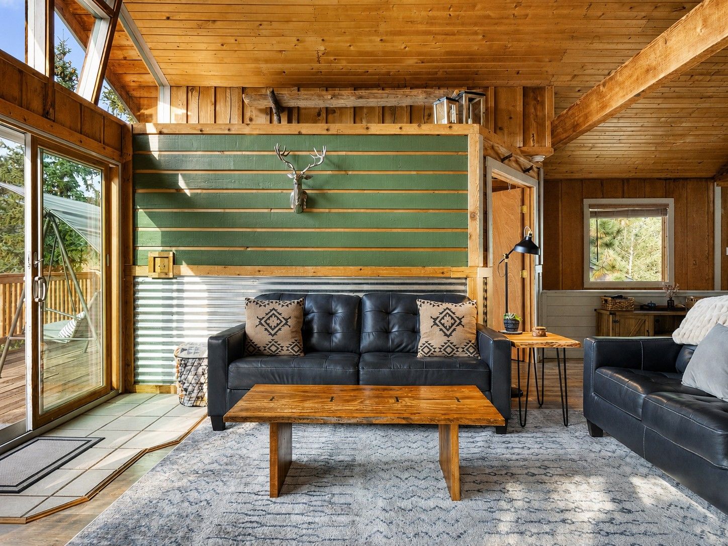 Interior acogedor de una cabaña con techo de madera, pared verde con tablones horizontales, sofá de cuero con cojines estampados, mesa de centro de madera y ventana con vistas a una terraza.