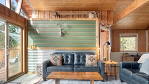 A cozy cabin interior with a wooden ceiling, a green wall with horizontal planks, a black leather sofa with patterned cushions, a wooden coffee table, and a window overlooking a deck.