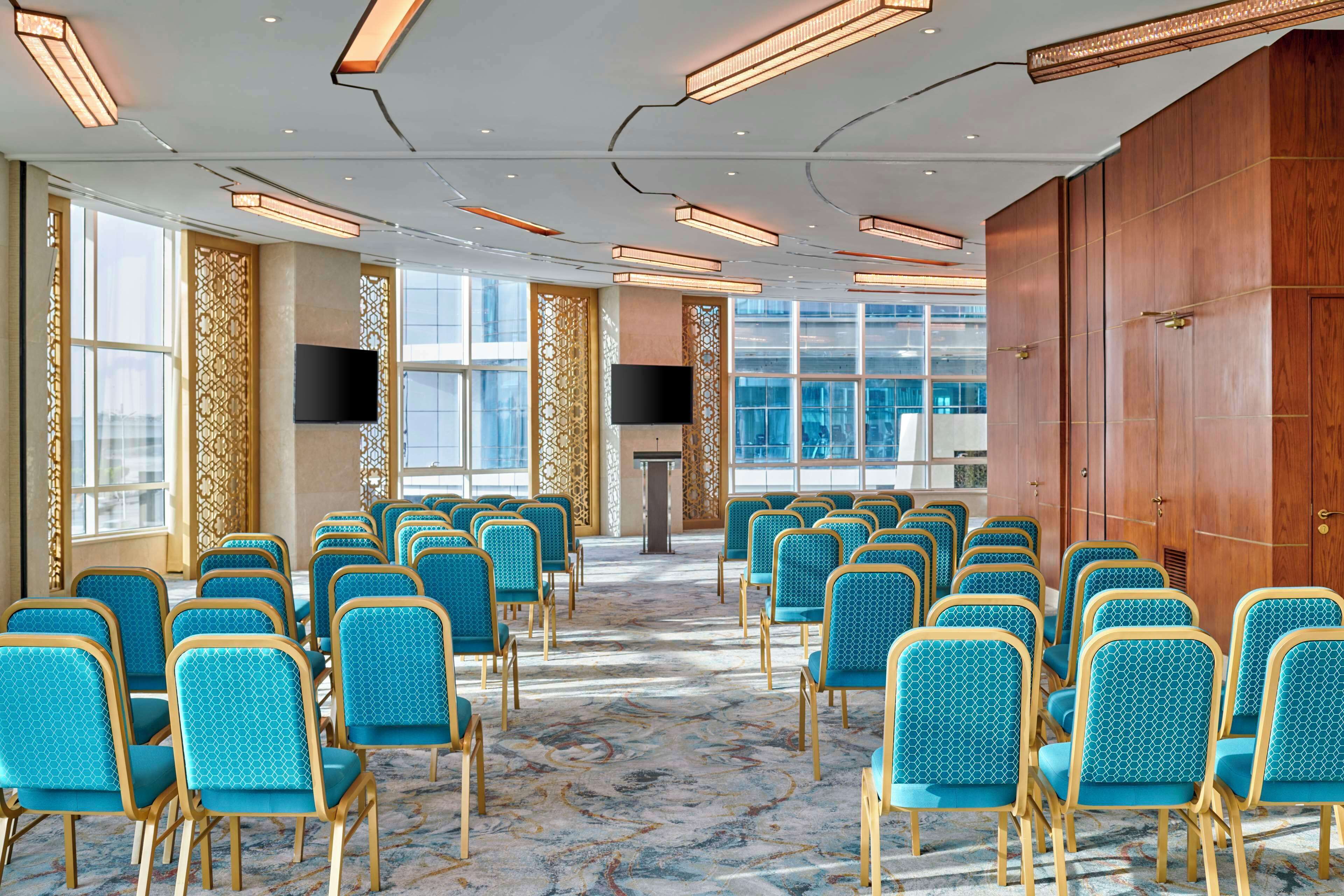 Un salón de conferencias con filas de sillas color turquesa, una pantalla grande y un panel de pared de madera.