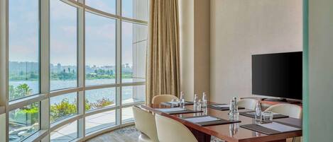 A conference room with a large window overlooking a river and cityscape, equipped with a flat-screen TV and a long table with chairs.
