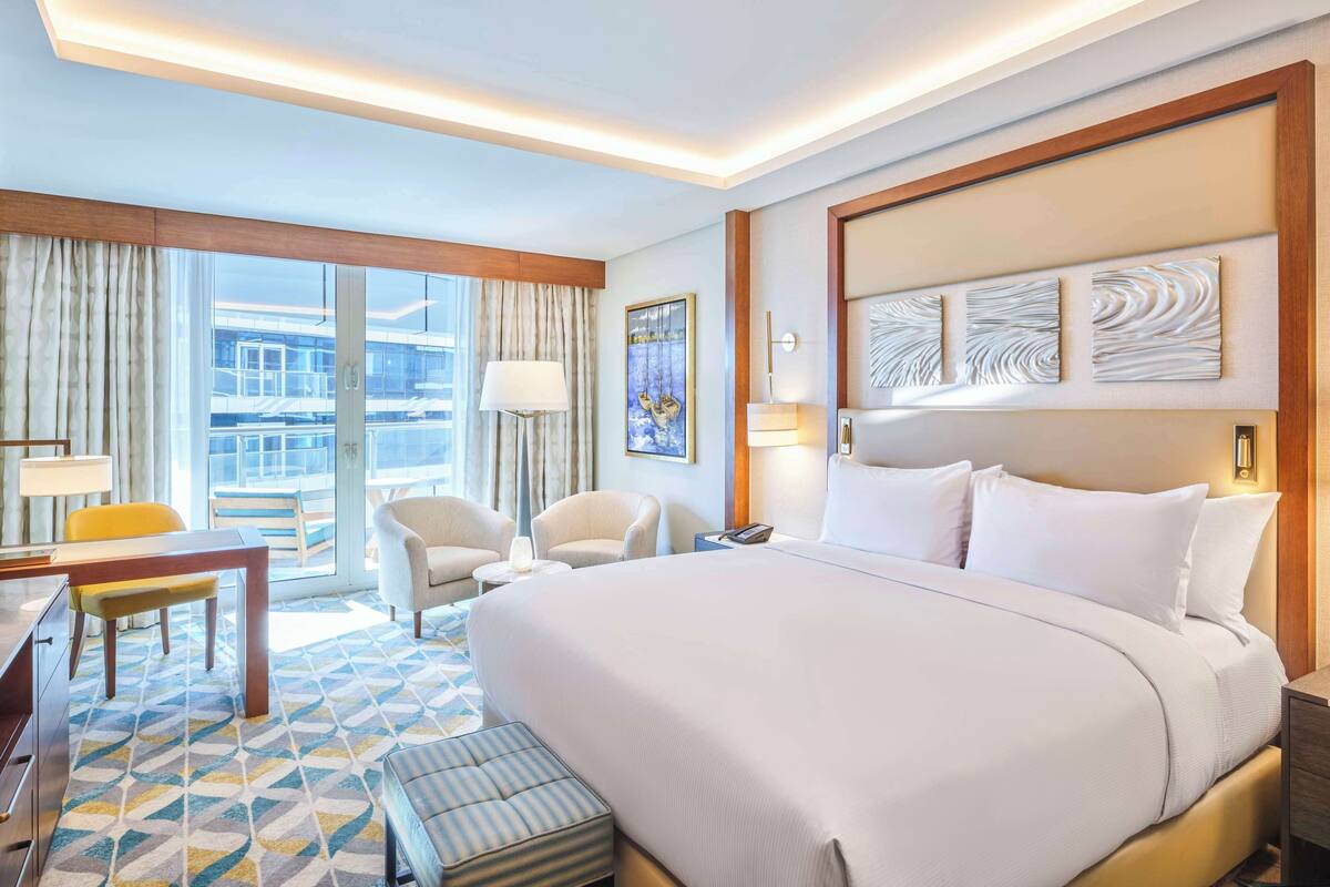 Une chambre d’hôtel avec un grand lit, deux fauteuils, un bureau avec une chaise, une petite table et une fenêtre avec des rideaux.