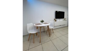 A modern, minimalist room with a white wall, a flat-screen TV mounted above a white console, and a small table with two white chairs. - Brand New Modern Studio with Pool (Luquillo)