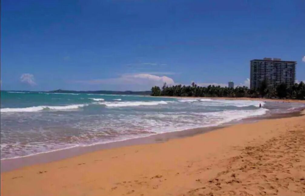 Une plage avec un ciel dégagé, des vagues sur l’océan et un grand immeuble en arrière-plan.