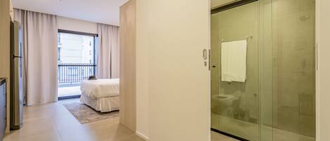A modern hotel room with a balcony, a glass-enclosed shower, and a bed with a white duvet.