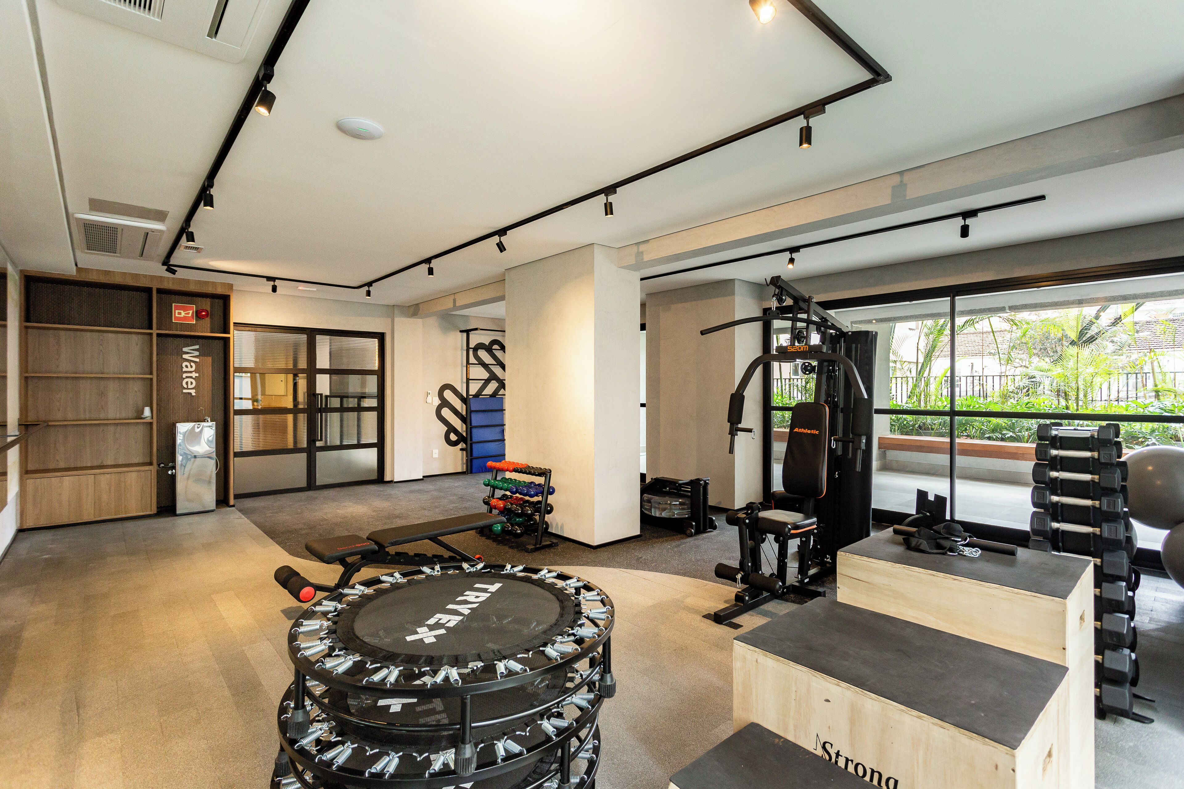A modern gym with various exercise equipment, a wooden bench, and a large window offering a view of greenery.