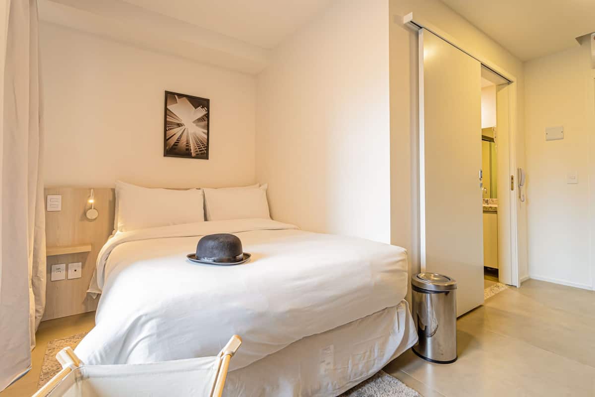Une chambre d’hôtel avec un lit, un tableau au mur, une poubelle et une porte donnant sur une autre pièce.