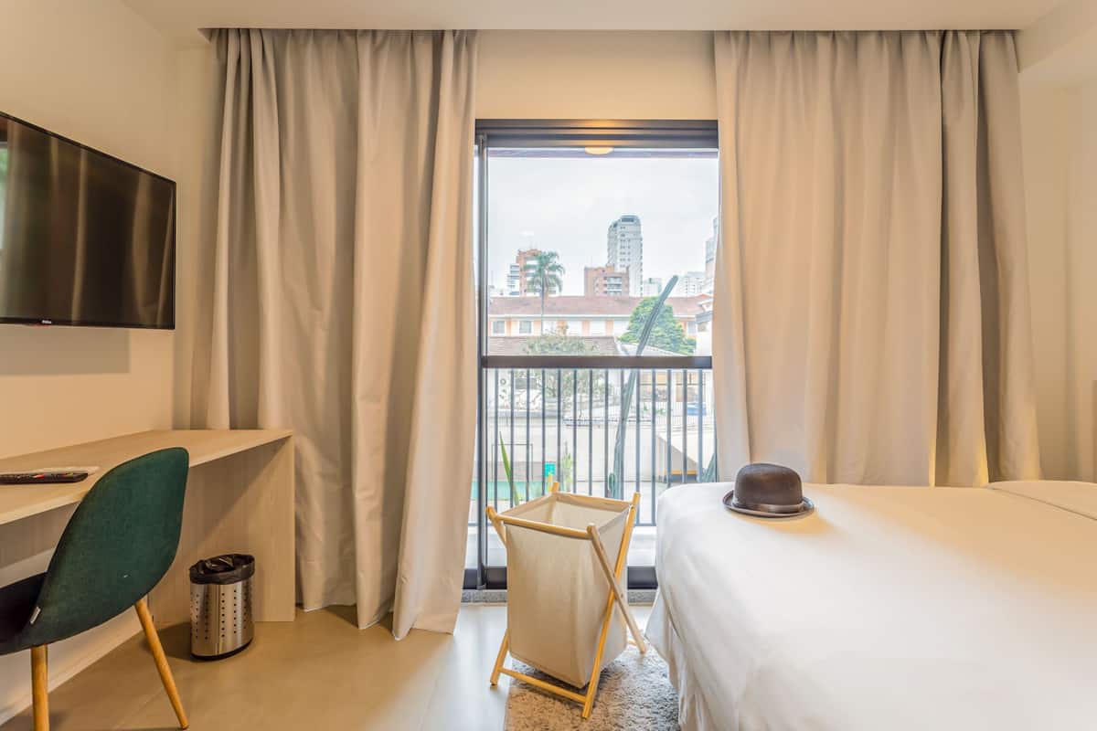 Une chambre d’hôtel avec un lit, un bureau, une chaise, une télévision et une vue sur les bâtiments et un palmier à l’extérieur.
