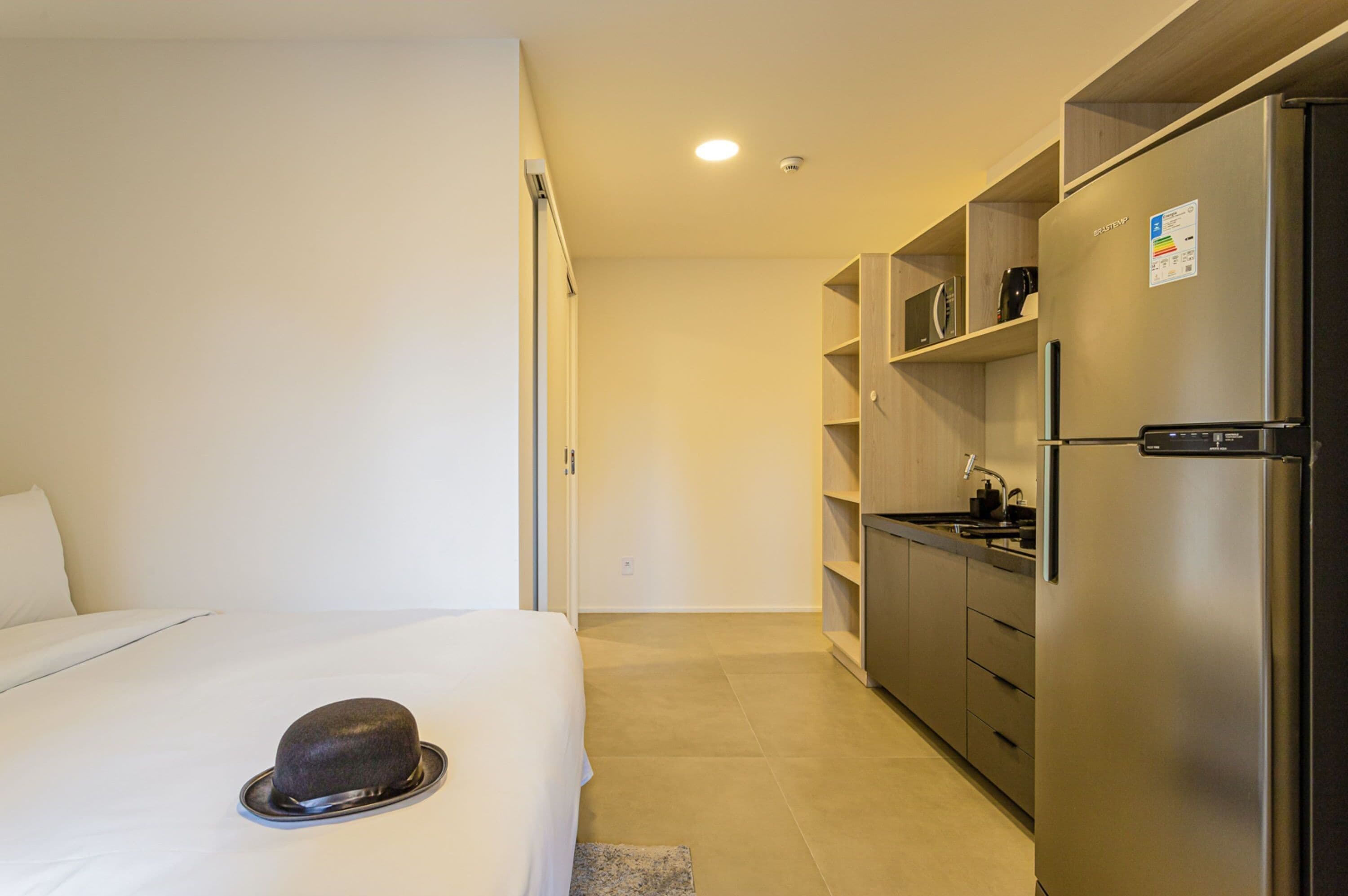 A modern hotel room with a bed, a kitchen area with a refrigerator, and a small countertop.