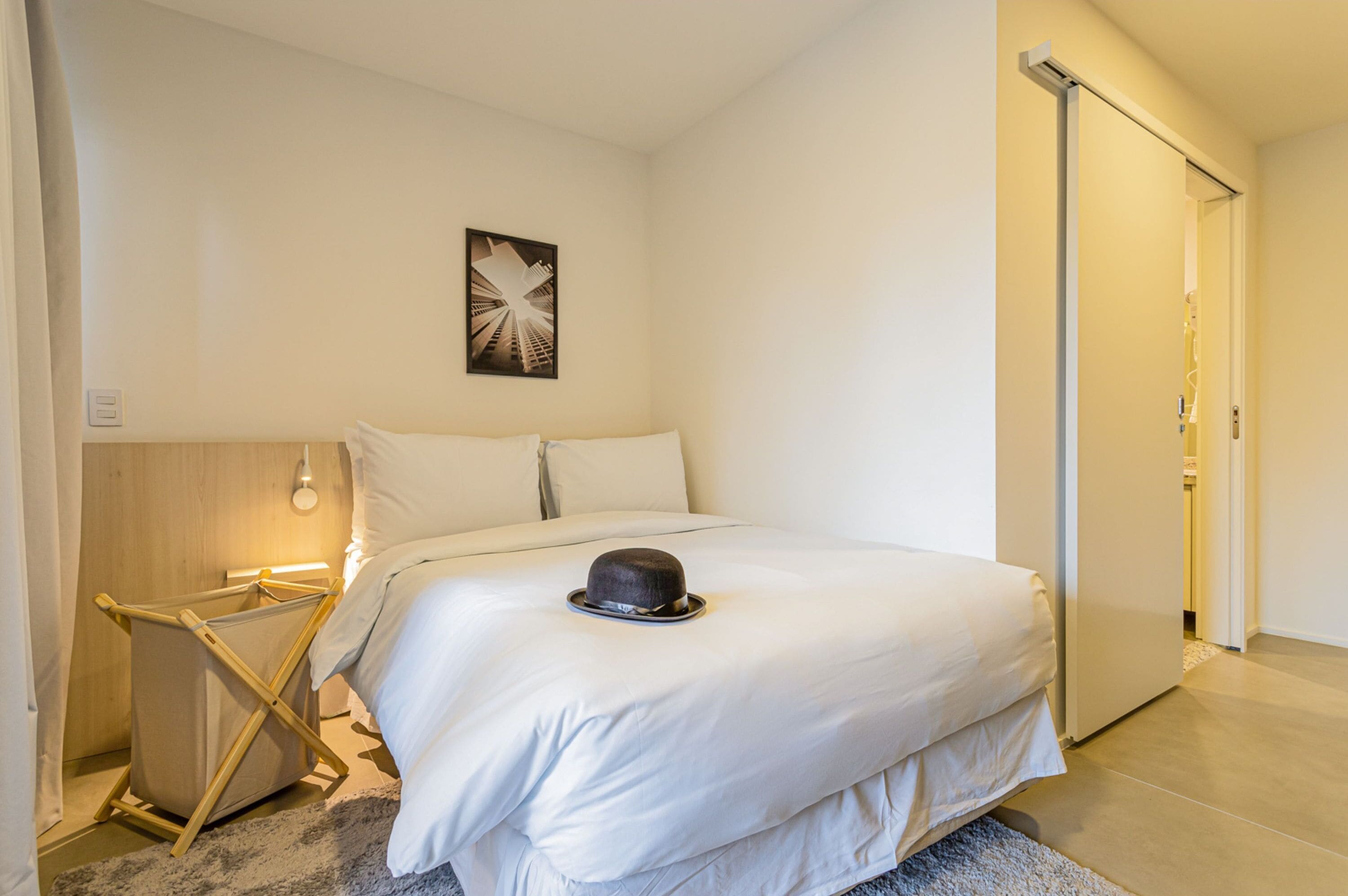 Una cama bien hecha con un sombrero encima, una mesita de noche de madera, un cuadro colgado en la pared y una puerta corredera.