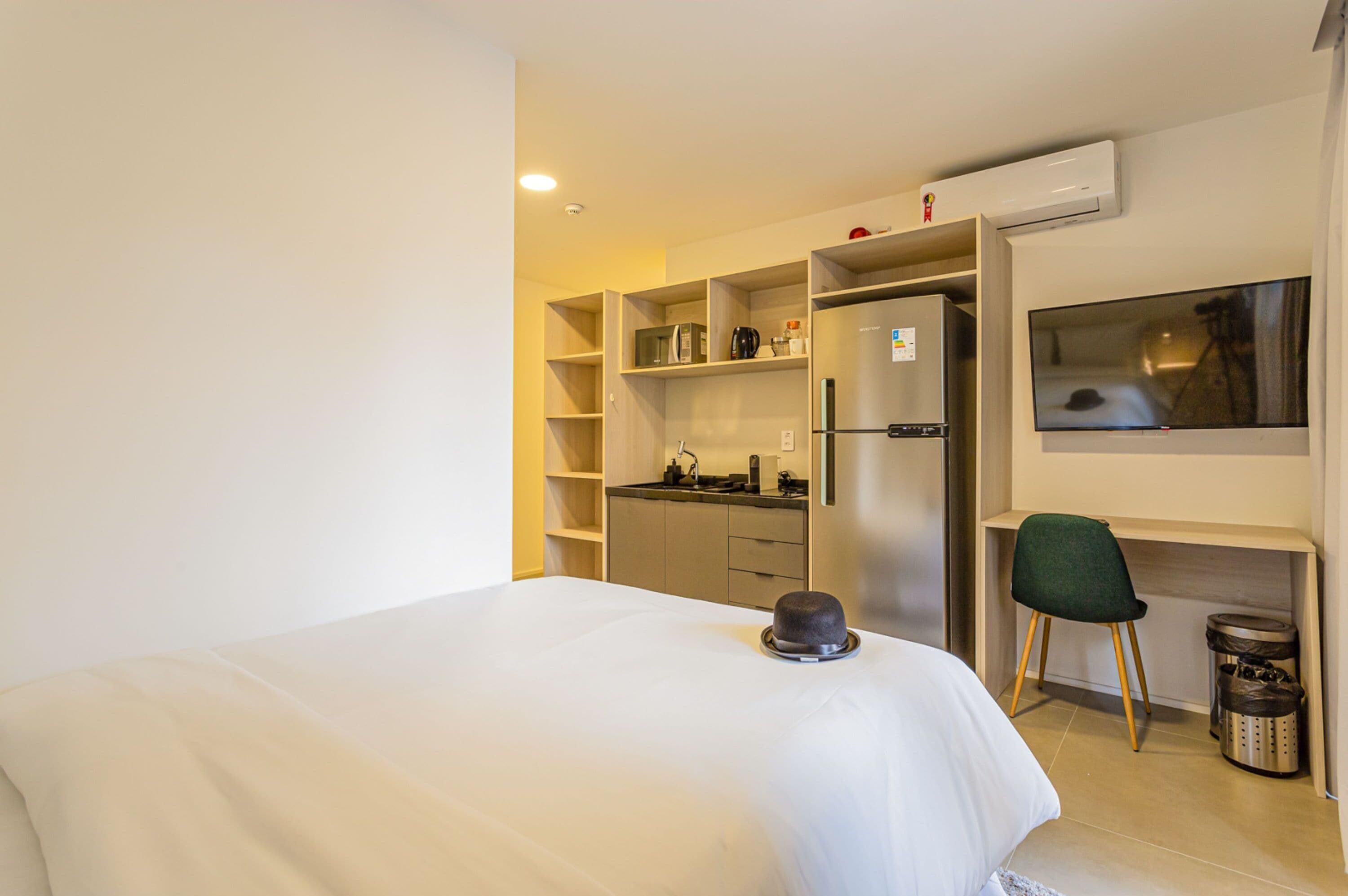A hotel room with a bed, a desk with a chair, a kitchenette, and a TV mounted on the wall.