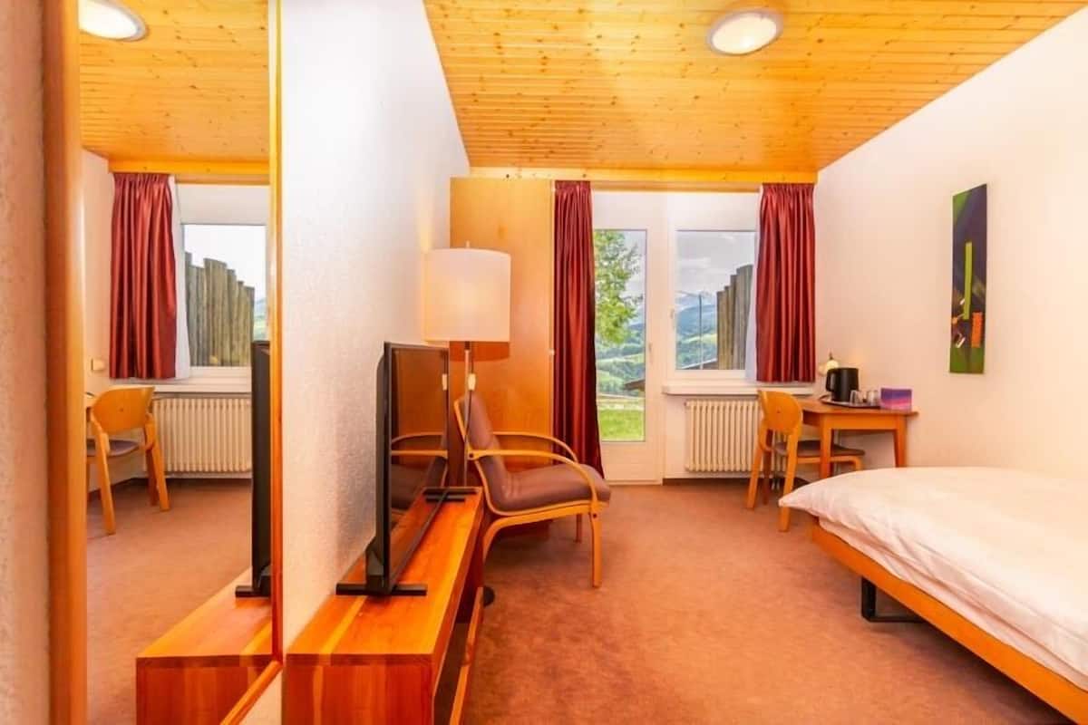 Ein Hotelzimmer mit Holzbalkendecke, einem Bett, einem Schreibtisch mit Stuhl, einem Fernseher und einem Fenster mit Vorhängen.