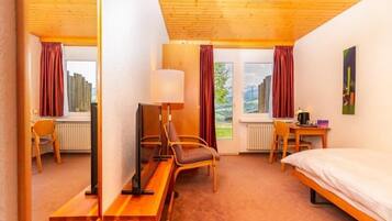 A hotel room with a wooden ceiling, a bed, a desk with a chair, a TV, and a window with curtains.