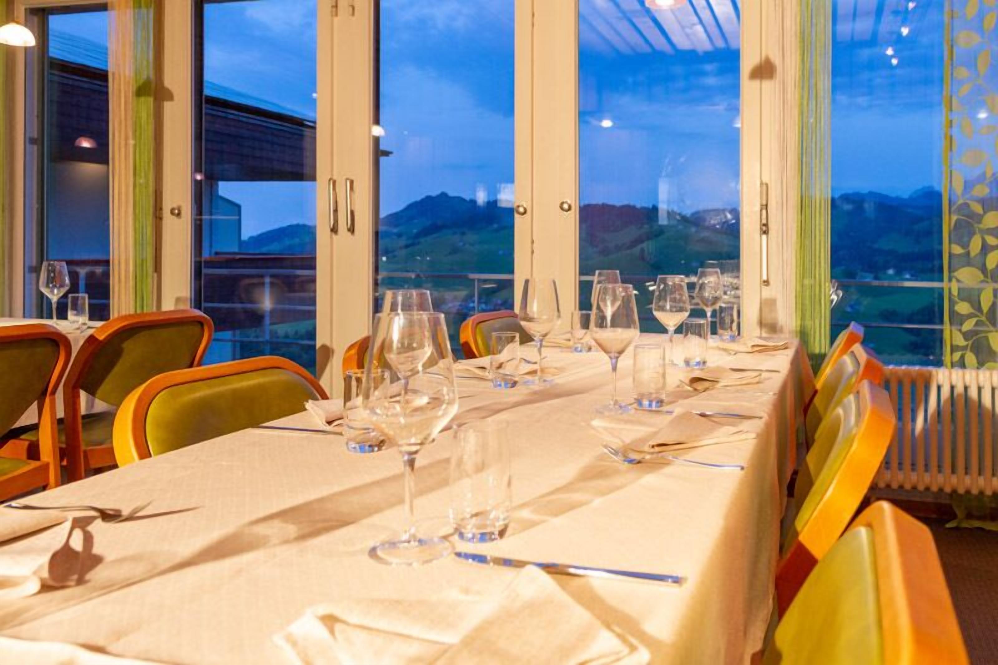 Mesa de comedor lista para una comida, con sillas, ventanas amplias y vistas al exterior.