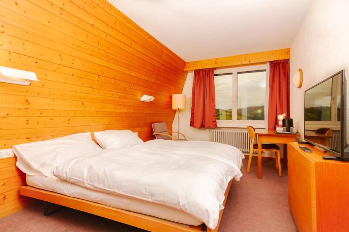 Ein Hotelzimmer mit holzgetäfelter, schräger Decke, einem Bett, einem Schreibtisch mit Stuhl, einem Fernseher und einem Fenster mit Vorhängen.