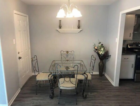 A dining area with a table and chairs, a chandelier, a vase with flowers, and a kitchen in the background.