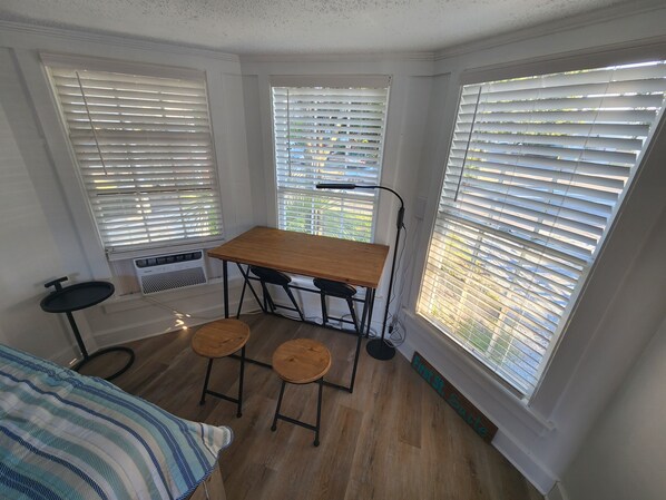 A compact room with a wooden table and two stools, large windows with blinds, a small round table, and a bed with a striped blanket.
