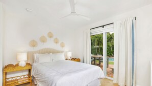 Quarto com uma cama grande, ventoinha no teto, mesa de cabeceira de vime e vista para uma piscina e vegetação lá fora.