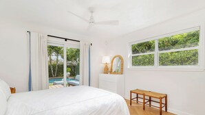 Quarto com cama, um banco de madeira, janela com vista para o verde, ventilador de teto e uma pequena mesa com abajur.