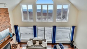 A cozy living room with a fireplace, a sofa, and a view of a snowy landscape through large windows. - Stoweic Views  - Luxury Dream Vacation Home (Stowe)