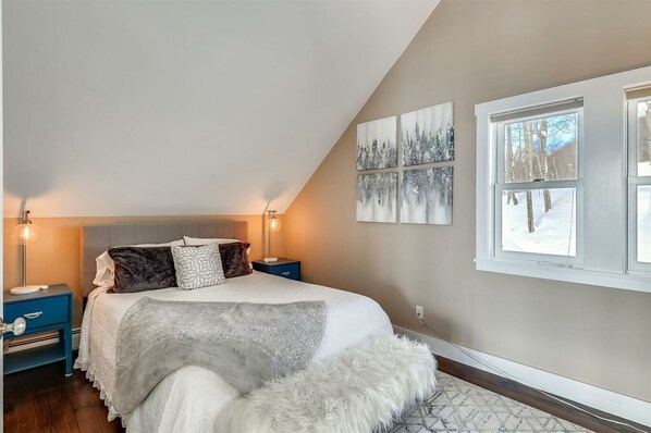 A bedroom with a sloped ceiling, a bed with a white and gray blanket, two bedside tables with lamps, and a window with a snowy view.
